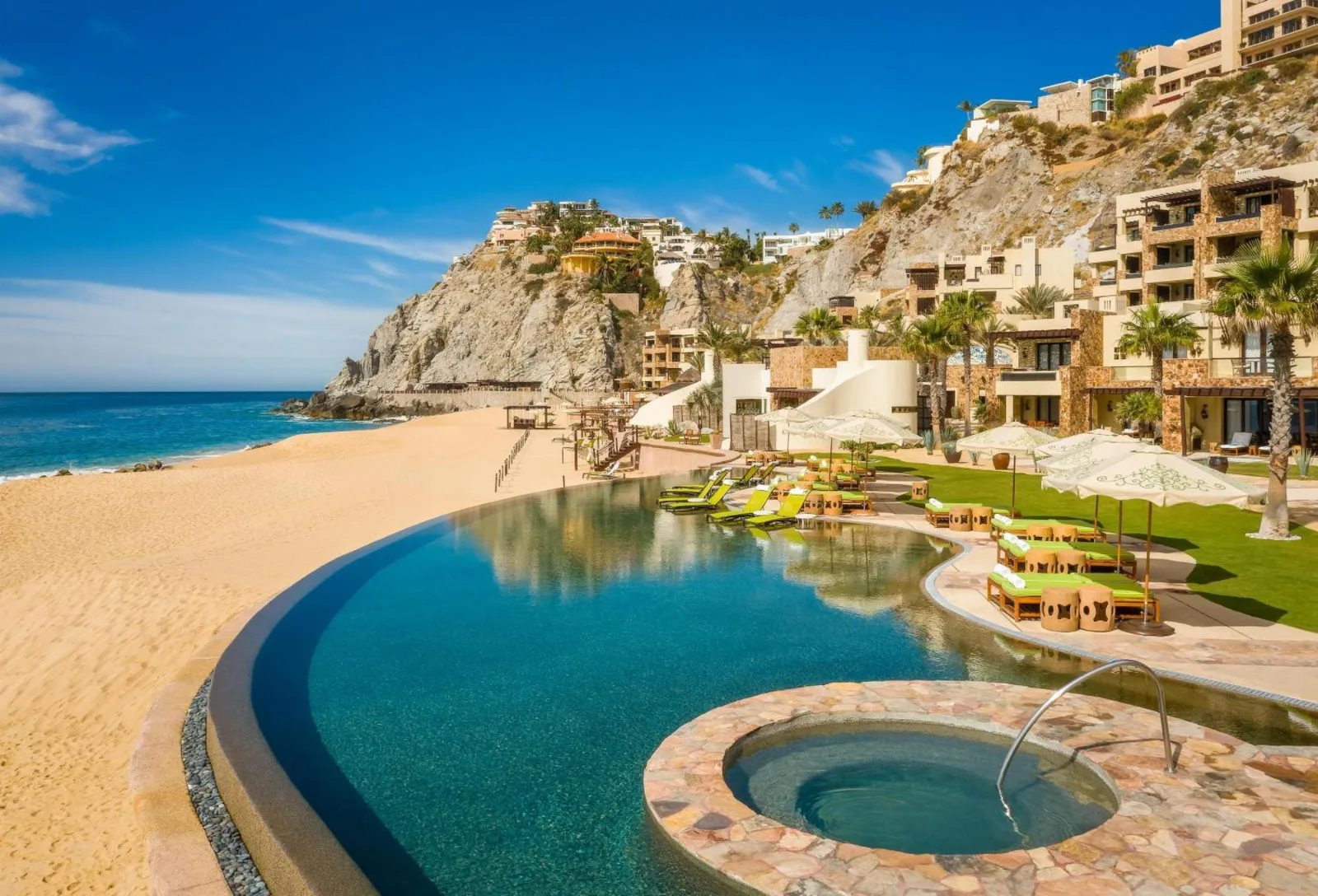 A curved infinity pool and stone hot tub overlook a sandy beach and cliffside resort buildings in Los Cabos, Mexico.
