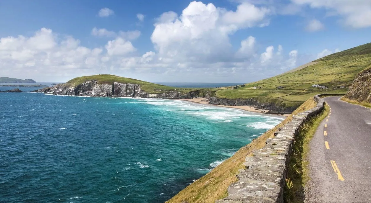 A paved coastal road with a stone wall winds along green hills overlooking a sandy cove and turquoise ocean in Ireland.